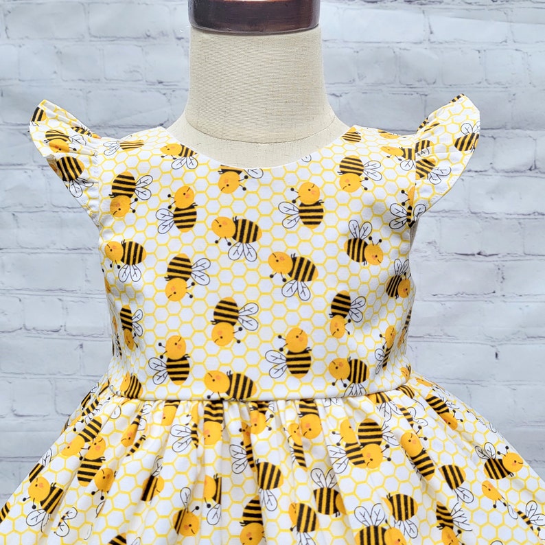 May include: A white dress with a yellow and black bee pattern. The dress has a ruffled neckline and short sleeves. The dress is perfect for a summer party or a day at the park.
