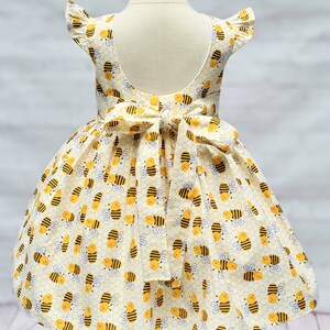 May include: A yellow and white dress with a bee pattern. The dress has a ruffled neckline and a bow at the back.