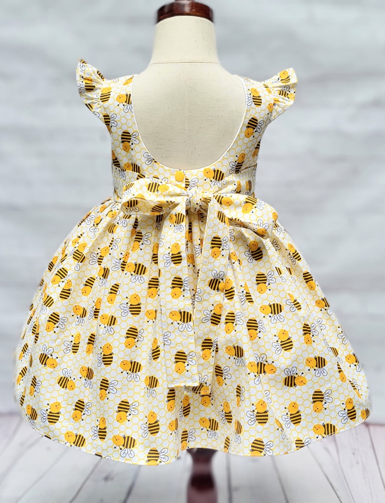 May include: A yellow and white dress with a bee pattern. The dress has a ruffled neckline and a bow at the back.