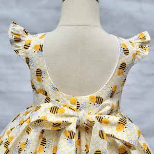 May include: A white dress with a yellow and black bee print. The dress has a scoop back and a bow tie at the waist.