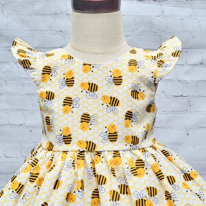 May include: A white dress with a yellow and black bee pattern. The dress has a ruffled neckline and short sleeves. The dress is perfect for a summer party or a day at the park.