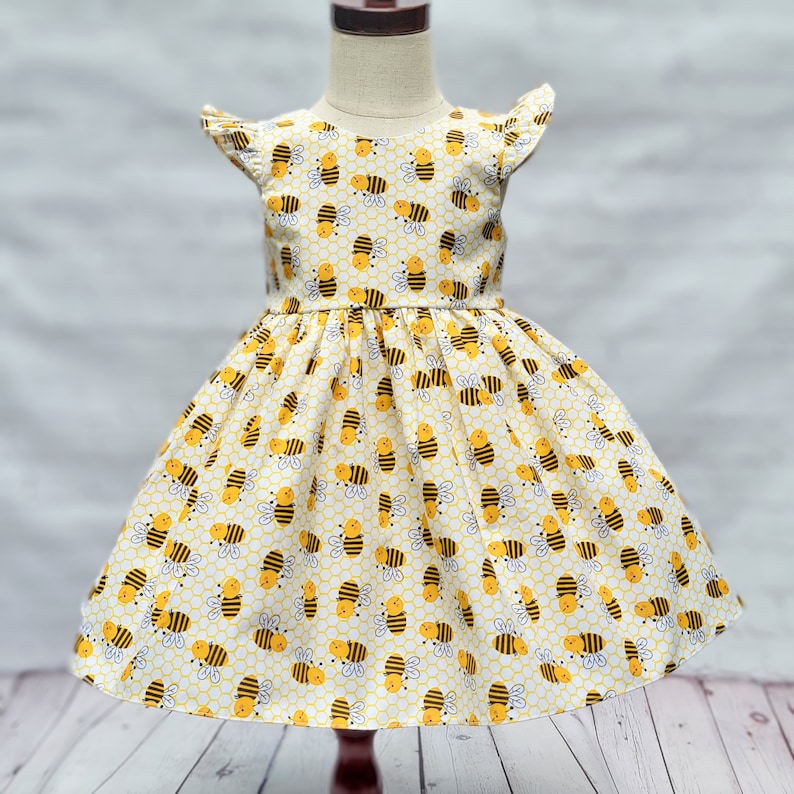 May include: A yellow and white dress with a bee pattern. The dress has short sleeves with ruffles and a full skirt.