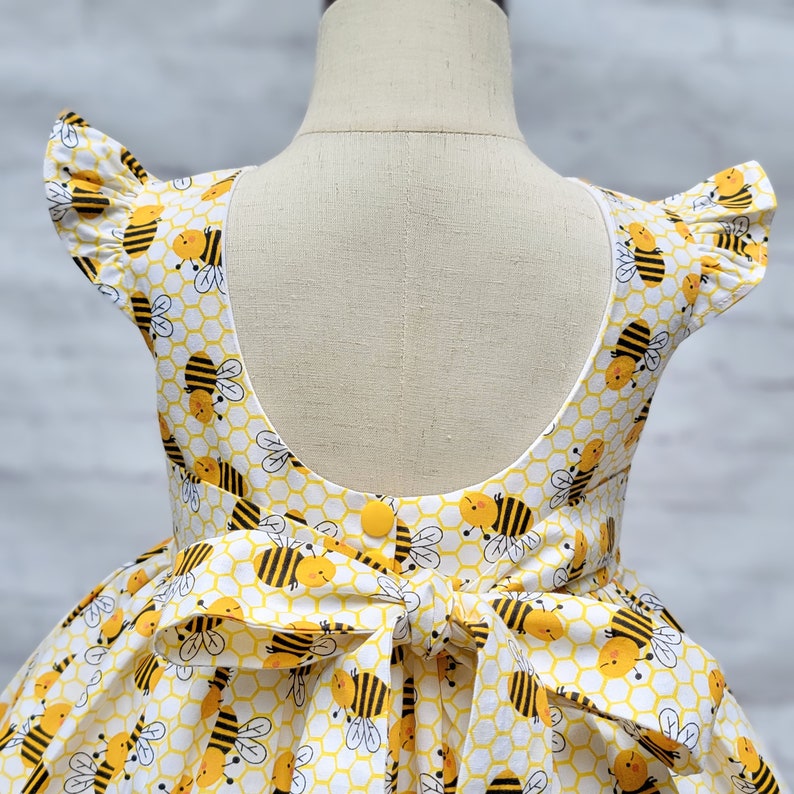 May include: A white dress with a yellow and black bee print. The dress has a scoop back and a bow tie in the back. The dress has short sleeves with ruffles.