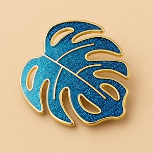 May include: A gold-toned metal brooch shaped like a monstera leaf. The leaf features cut-out sections and is filled with a vibrant, glittery blue enamel finish. The edges of the leaf and the internal veins are outlined in a polished gold metal.
