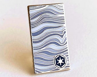 May include: A rectangular enamel pin featuring a pattern of wavy lines in shades of light blue, grey, and white. In the bottom right corner, there is a circular dark blue emblem with a white six-spoked star symbol surrounded by a thin white border.