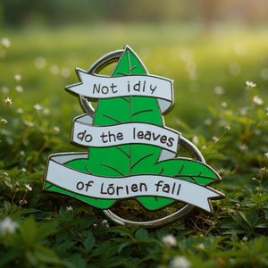 May include: A green enamel pin shaped like a leaf, wrapped in a white ribbon with the black text "Not idly do the leaves of Lórien fall." The pin features a metallic silver border and a circular backing element, presented in a clear, sharp design.