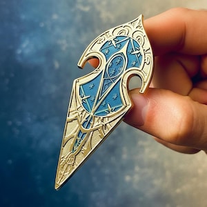 May include: A gold-toned metal enamel pin shaped like a pointed, elongated shield. The design features intricate gold filigree patterns over a matte blue background, accented with small, embossed star shapes throughout the centre.