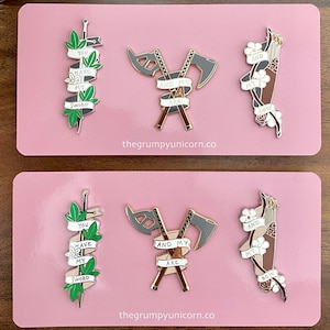May include: Two sets of enamel pins featuring a sword, axe, and bow and arrow. Each pin has a banner with the text "You have my sword", "And my axe", and "And my bow". The pins are on a pink background.