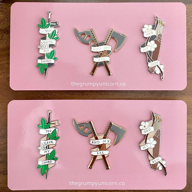 May include: Two sets of enamel pins featuring a sword, axe, and bow and arrow. Each pin has a banner with the text "You have my sword", "And my axe", and "And my bow". The pins are on a pink background.