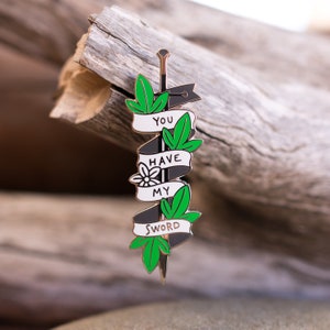 May include: A black and white enamel pin shaped like a sword with green leaves and the text "You Have My Sword" on it.