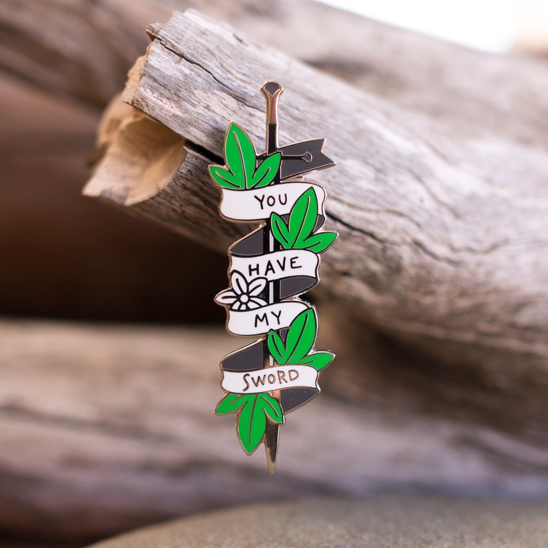 May include: A black and white enamel pin shaped like a sword with green leaves and the text "You Have My Sword" on it.