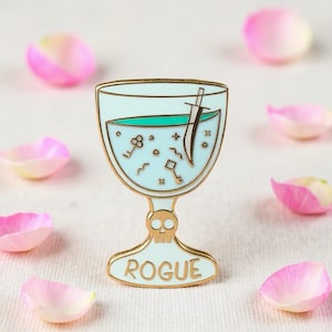 May include: A gold-colored enamel pin in the shape of a goblet, featuring a light blue liquid with a sword and keys. The base of the goblet has a skull design and the word "ROGUE". Pink and white flower petals are scattered around the pin.