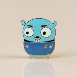 Pode incluir: Um alfinete de esmalte azul com um personagem de desenho animado com uma expressão mal-humorada. O personagem tem olhos brancos grandes, um nariz pequeno e uma roupa azul com as letras "A" e "D". O alfinete tem uma borda dourada.