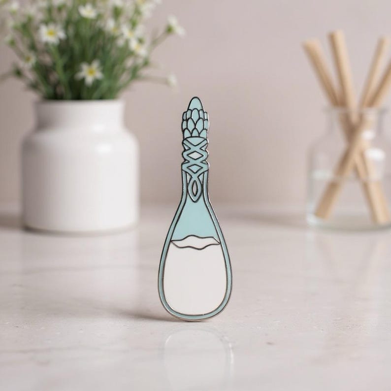 K&ouml;nnte beinhalten: Ein Metall-Emaille-Pin in Form einer Zaubertrankflasche mit einem hellblauen oberen Teil und einem wei&szlig;en unteren Teil. Der Flaschenhals weist ein detailliertes, filigranes silberfarbenes Gittermuster auf, w&auml;hrend der gesamte Pin von einem d&uuml;nnen silbernen Metallrand umgeben ist.