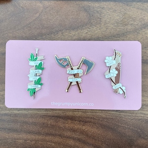 May include: Three enamel pins on a pink background. The pins are of a sword, an axe and a bow and arrow. The sword pin has the text "You have my sword". The axe pin has the text "And my axe". The bow and arrow pin has the text "Oh and my bow".