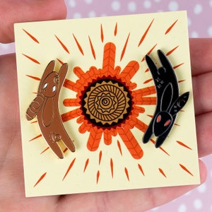 May include: Two enamel pins depicting stylized rabbits. One is brown with gold detailing, the other is black with red eyes. They are set against a yellow background with an orange sunburst design. The pins are suitable for a jacket, bag, or other accessory.