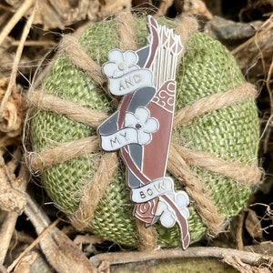 May include: A silver enamel pin with a brown and black bow and arrow, with white flowers and the words "My Bow And" on it.