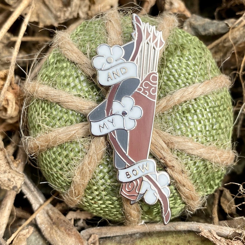 May include: A silver enamel pin with a brown and black bow and arrow, with white flowers and the words "My Bow And" on it.
