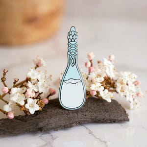 K&ouml;nnte beinhalten: Eine Metall-Emaille-Anstecknadel in Form einer hohen, schlanken Flasche mit einem hellblauen oberen Teil und einem wei&szlig;en unteren Teil. Der Flaschenhals zeigt ein dekoratives, symmetrisches Gittermuster in Schwarz, getrennt durch eine feine Linie.