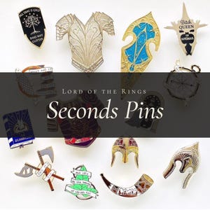 May include: A collection of metal enamel pins featuring various designs including a shield with a tree, armour, a blue and gold pointed shape, a crown, a book, axes, a leaf, a horn, and helmets, with some pins displaying text.