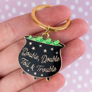 May include: A black cauldron keychain with gold trim and a gold keyring. The cauldron is filled with green bubbles and has the words "Double, Double Toil & Trouble" in gold lettering. The background is a light pink with white polka dots.