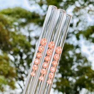 Pu&ograve; includere: Set di tre cannucce da bere in vetro con un design geometrico rosa e bianco. Le cannucce sono trasparenti e hanno un design rosa e bianco a forma di dadi.