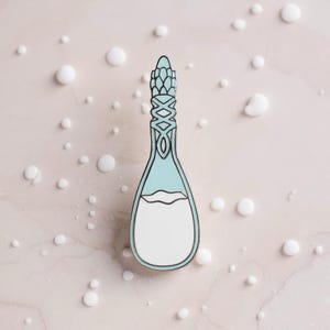 K&ouml;nnte beinhalten: Eine Metall-Emaille-Anstecknadel in Flaschenform mit einem hellblauen oberen Teil und einem wei&szlig;en unteren Teil. Der Flaschenhals weist ein dekoratives, kompliziertes Gittermuster in Hellblau und Silber mit einer klaren Umrandung auf.