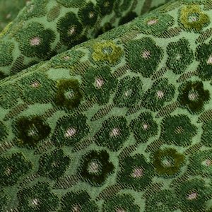 Op de afbeelding: Close-up van een groene stof met bloemenpatroon. De stof heeft een textuur met een herhalend patroon van donkergroene bloemen met lichtroze centra. De achtergrond is een lichtere tint groen.