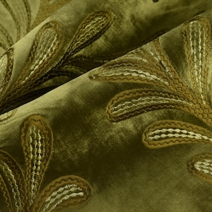 May include: Close-up of olive green fabric with embroidered leaf-like patterns in shades of gold and brown. The fabric is folded, showing the texture and detail of the design. The pattern is repeated across the surface.