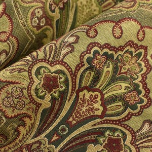 May include: Close-up of a folded fabric with a detailed paisley pattern. The fabric features a blend of olive green, burgundy, and gold colors. The design includes floral and scroll-like motifs, creating a textured and ornate appearance.