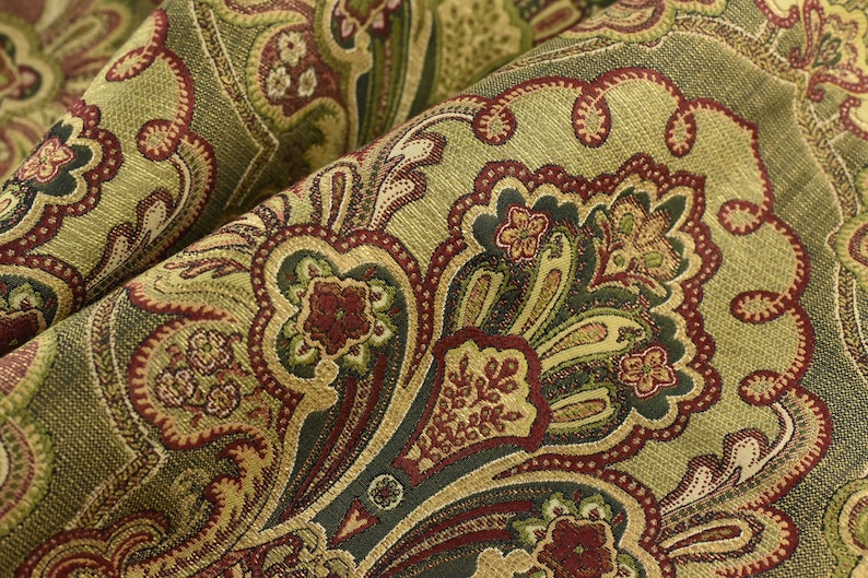 May include: Close-up of a folded fabric with a detailed paisley pattern. The fabric features a blend of olive green, burgundy, and gold colors. The design includes floral and scroll-like motifs, creating a textured and ornate appearance.