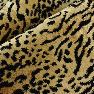 May include: Close-up of folded fabric featuring a tan base with a black leopard print design. The fabric has a soft, textured appearance. The pattern comprises irregular black spots and elongated shapes.