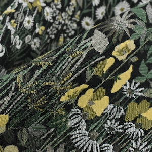 May include: Close-up of a black fabric with a floral pattern. The design features yellow, white, and gray flowers, along with green leaves and stems. The fabric has a textured appearance.