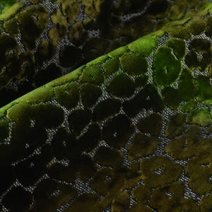 May include: Close-up of a luxurious fabric with a velvet texture. The fabric features a pattern of large, irregular shapes in shades of green and brown, creating a gradient effect. The background is a neutral gray.