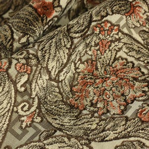 May include: Close-up of a fabric with a detailed floral pattern. The design features large, stylized flowers and foliage in shades of brown, beige, and coral. The fabric has a textured appearance, with a subtle geometric background.