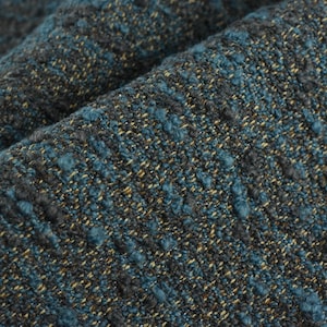 May include: Close-up of a textured fabric with a mix of teal, gray, and gold threads. The fabric has a nubby, woven appearance, with a slightly irregular surface. The folds in the fabric create shadows and depth.