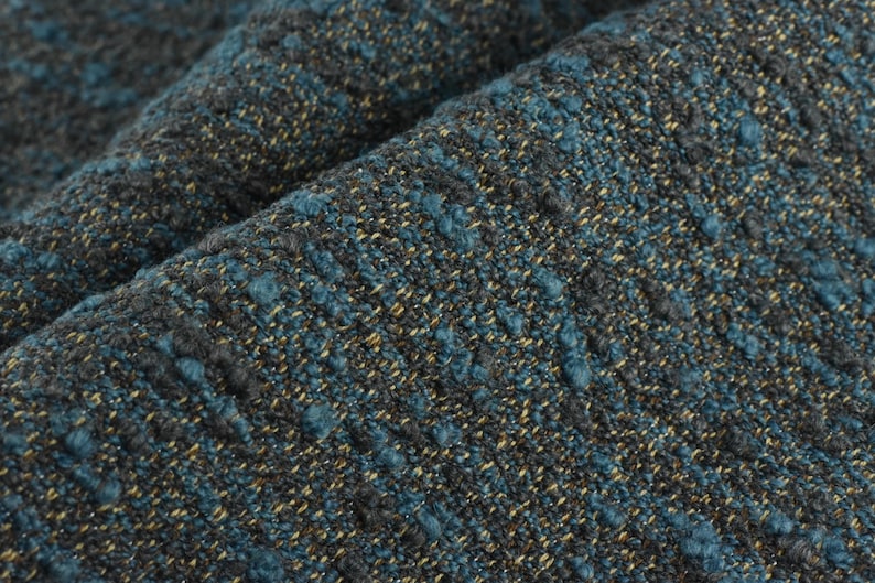 May include: Close-up of a textured fabric with a mix of teal, gray, and gold threads. The fabric has a nubby, woven appearance, with a slightly irregular surface. The folds in the fabric create shadows and depth.