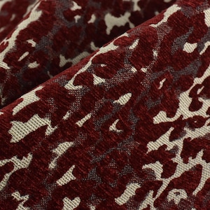 Può includere: Primo piano di un tessuto strutturato con un motivo ripetuto. Il tessuto presenta un colore bordeaux intenso con un design astratto color crema. Il tessuto è piegato, mostrando profondità e consistenza.