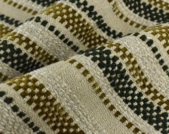 May include: Close-up of a woven fabric with a textured surface. The fabric features a repeating pattern of olive green and dark green squares and lines against a beige background. The fabric is folded, showing the texture and depth of the weave.