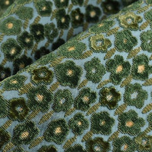 Könnte beinhalten: Nahaufnahme eines Stoffes mit floralem Muster. Der Stoff hat einen hellblauen Hintergrund mit einem sich wiederholenden Muster aus grünen Blumen und braunen Stielen. Die Blumen haben ein strukturiertes, samtartiges Aussehen.