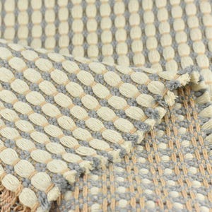 May include: Close-up of a woven rug with a textured pattern of beige and gray ovals. The rug has a fringe border.