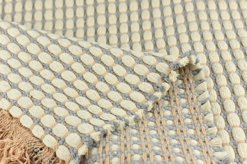 May include: Close-up of a woven rug with a textured pattern of beige and gray ovals. The rug has a fringe border.
