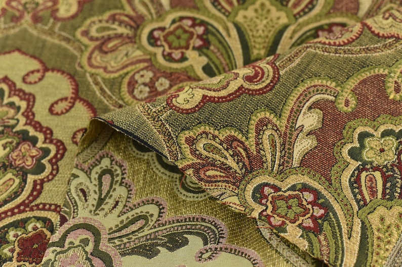 May include: Close-up of a folded fabric with a detailed paisley pattern. The fabric features a blend of olive green, burgundy, and gold colors. The design includes floral and curved elements, creating a vintage aesthetic.