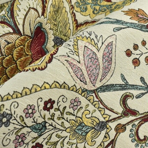 Op de afbeelding: Een close-up van een stof met een bloemenpatroon. De stof is crèmekleurig met een verscheidenheid aan bloemen in tinten rood, roze, geel en blauw. De bloemen zijn ingewikkeld en gedetailleerd, en de stof heeft een textuur.