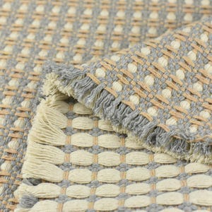 May include: A woven rug with a gray and beige pattern. The rug has a fringe border and a textured surface.