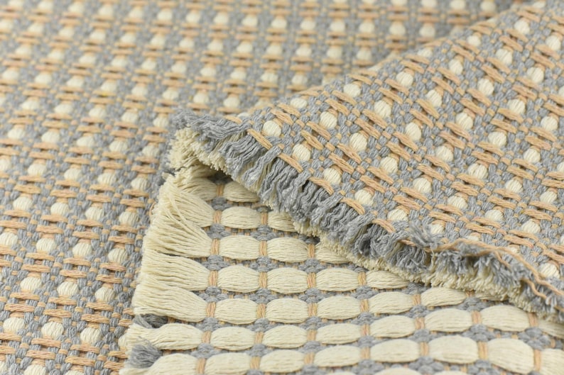 May include: A woven rug with a gray and beige pattern. The rug has a fringe border and a textured surface.
