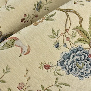 May include: A cream-colored fabric featuring a woven pattern of colorful birds, blooming flowers in shades of blue and red, and green foliage. The textile displays a detailed botanical and avian design with textured threads throughout the surface.