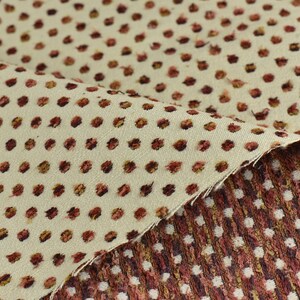 May include: Close-up of a beige fabric with a repeating pattern of small, brown polka dots. The fabric is folded over, revealing a second layer of fabric with a similar pattern but in a darker brown colour.