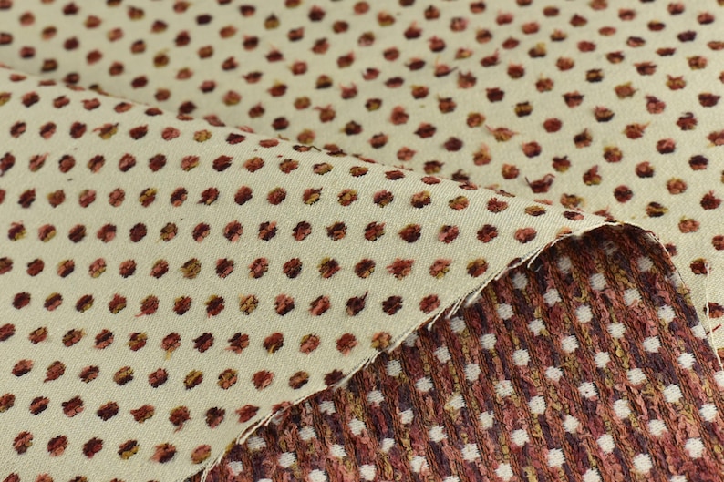 May include: Close-up of a beige fabric with a repeating pattern of small, brown polka dots. The fabric is folded over, revealing a second layer of fabric with a similar pattern but in a darker brown colour.