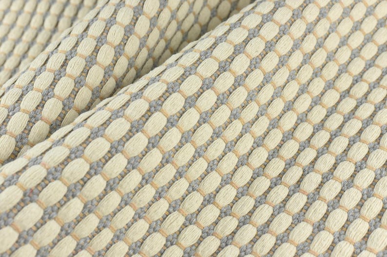 May include: Close-up of a woven fabric with a textured pattern. The fabric is a light beige color with a subtle gray and light brown pattern.